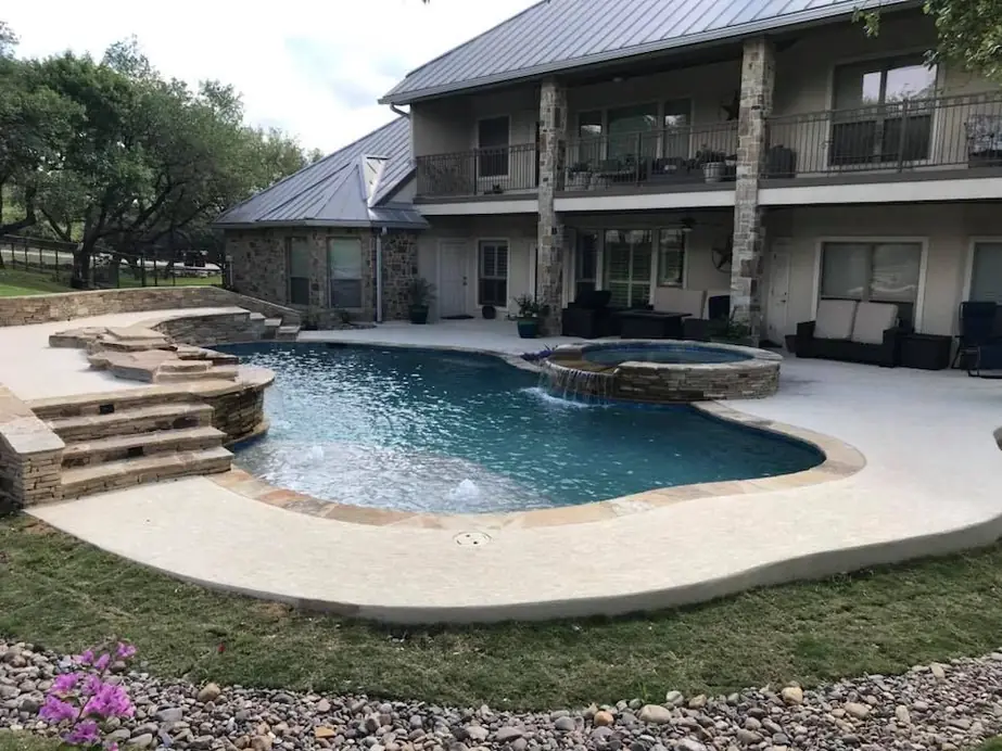 Freeform gunite pool with a raised stone spa and stone steps behind a two-storey Hill Country home