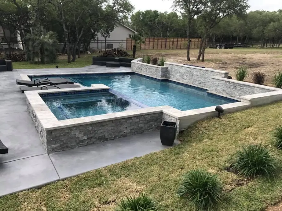 Rectangular pool and raised spa wrapped in stacked stone beside a young lawn